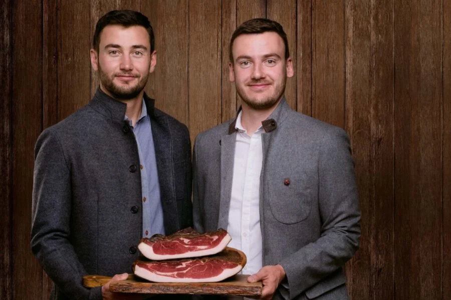 Hersteller der Recla halten Brett mit Südtiroler Speck g.g.A. vor Holzwand und blicken in die Kamera