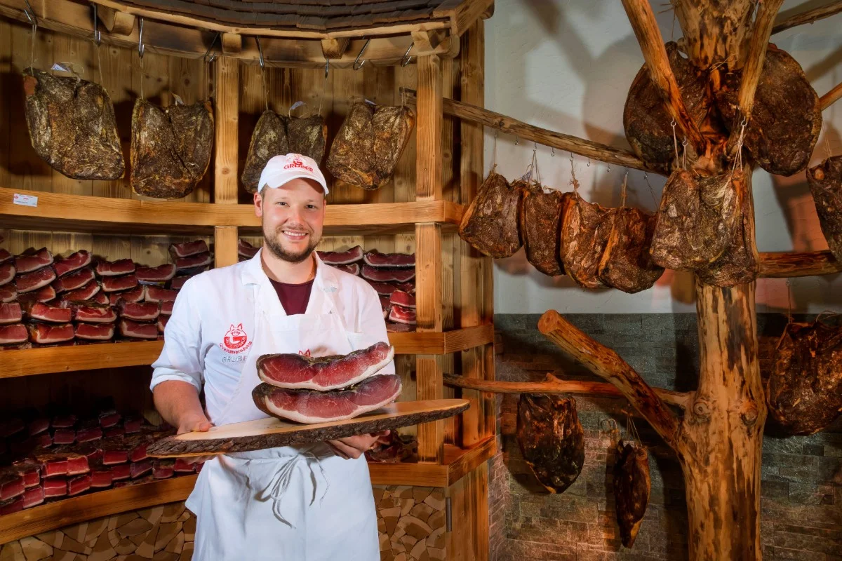 Werner Gruber hält Brett mit Südtiroler Speck g.g.A. im Geschäft, im Hintergrund hängen mehrere Stücke