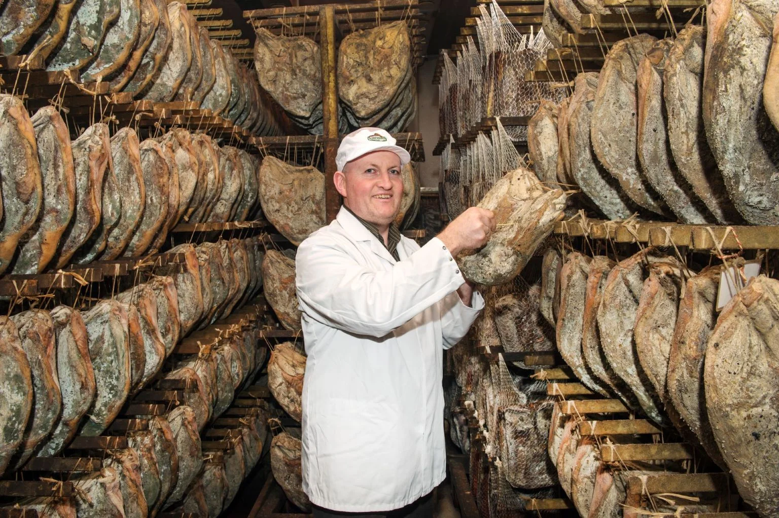 Günther Windegger holding a piece of Speck Alto Adige PGI in a curing room with hanging speck
