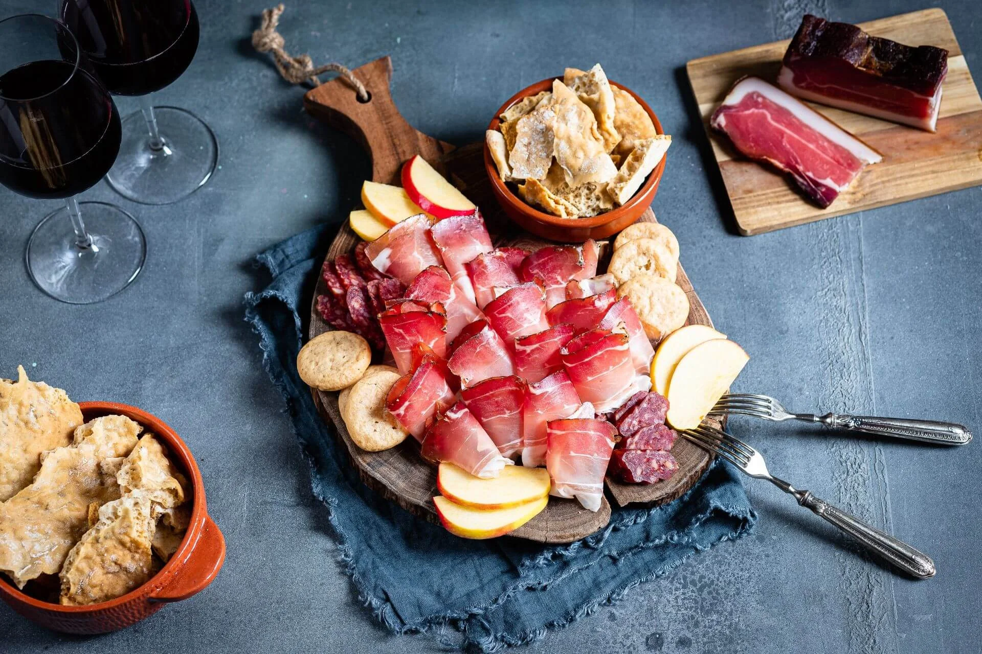 Holzbrett mit Scheiben Südtiroler Speck g.g.A., Äpfeln und Schüttelbrot auf rustikalem Tisch mit Rotweingläsern.