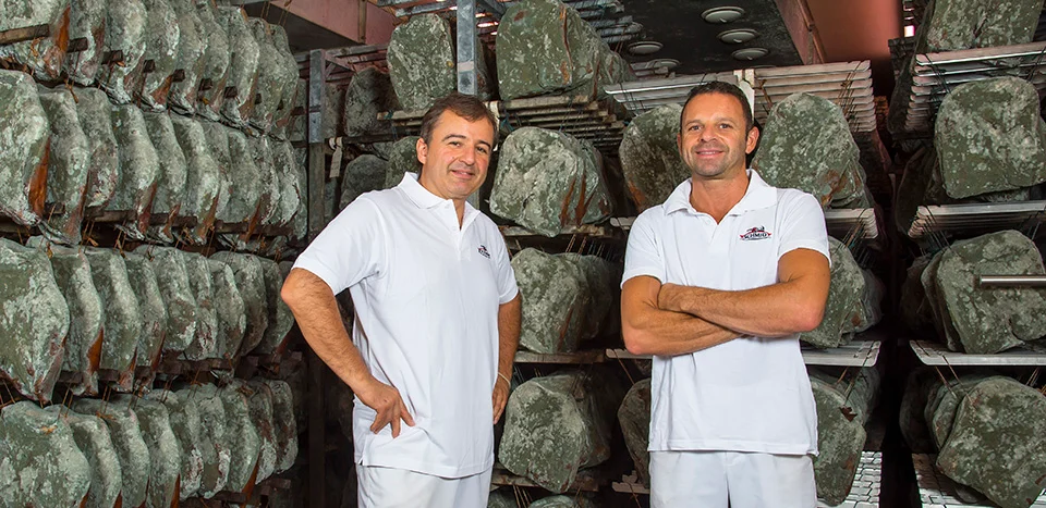 Thomas and Joachim Schmid posing in front of shelves with Speck Alto Adige PGI in a curing room