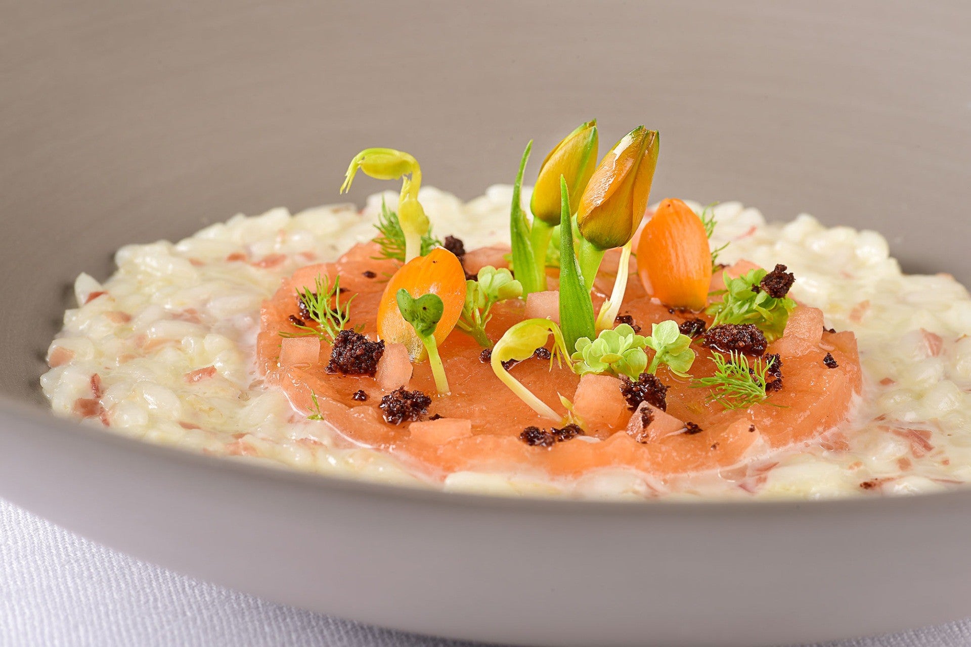 Risotto crémeux au speck et melon de Mantoue, garni d’herbes et de fleurs comestibles, dressé avec élégance.