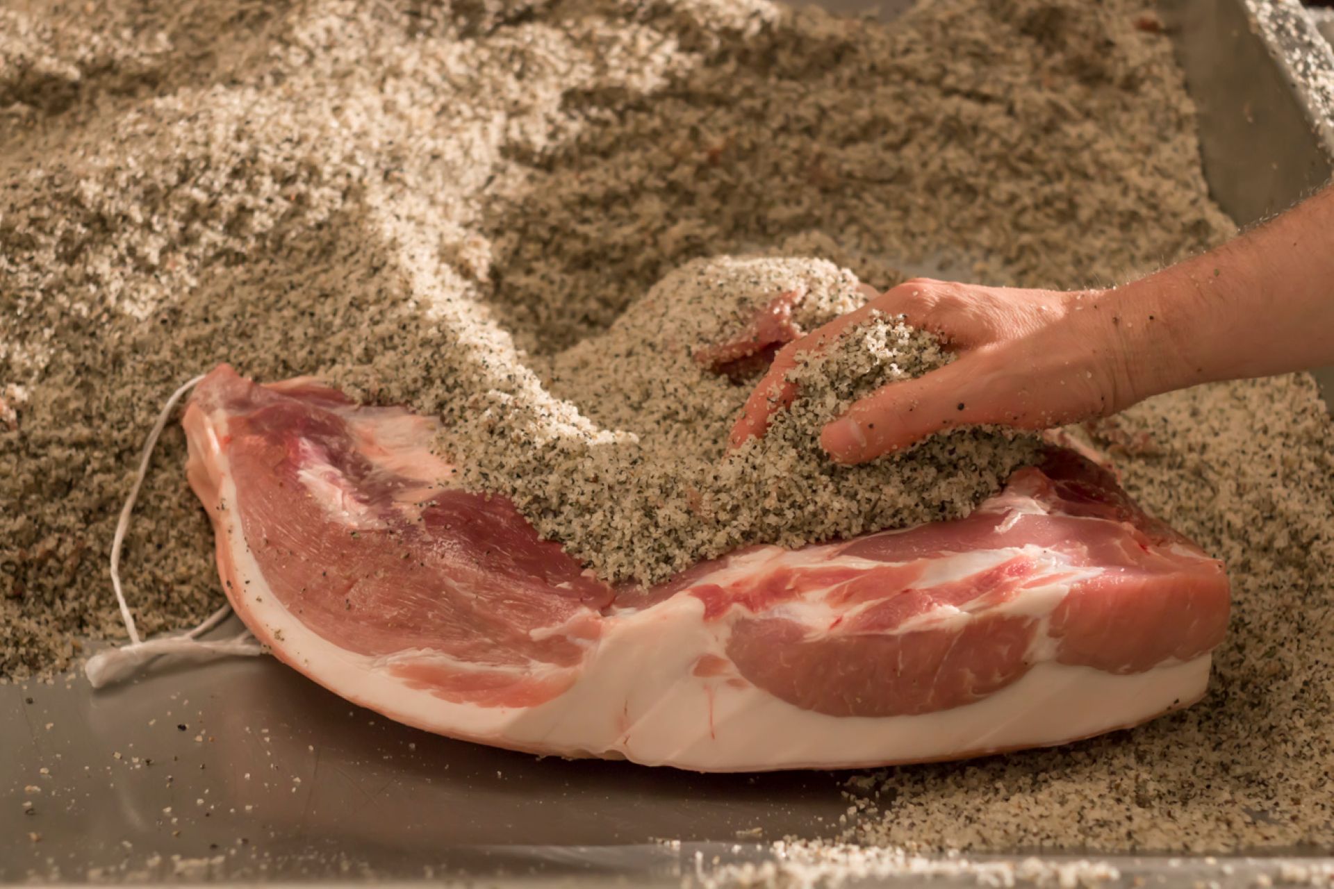 Hand bestreut Gewürze auf ein Stück Südtiroler Speck g.g.A. während der Verarbeitung.