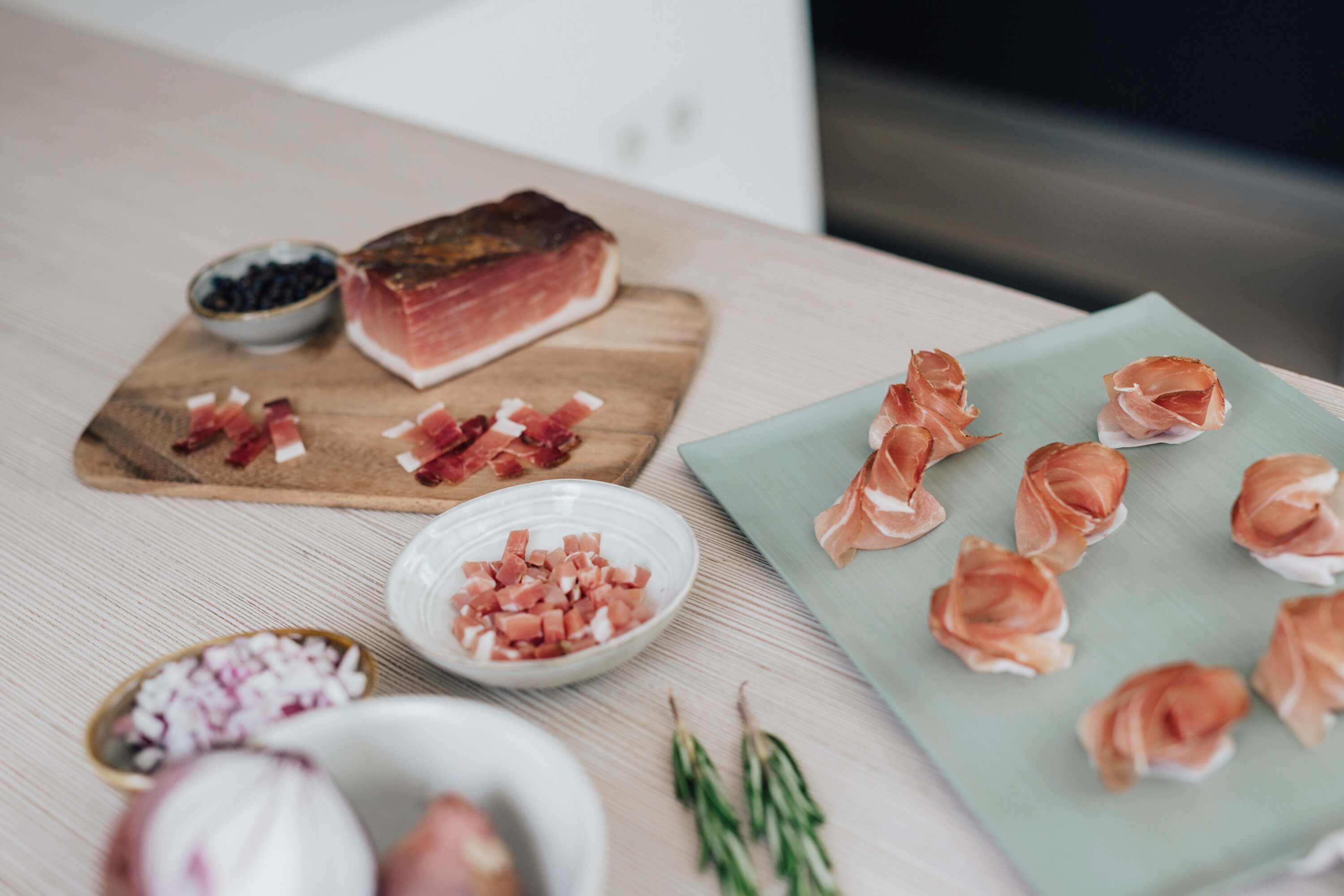 Scheiben Südtiroler Speck g.g.A. auf Teller, daneben Stück und verschiedene Schnitte auf Brett mit Zwiebeln und Kräutern.