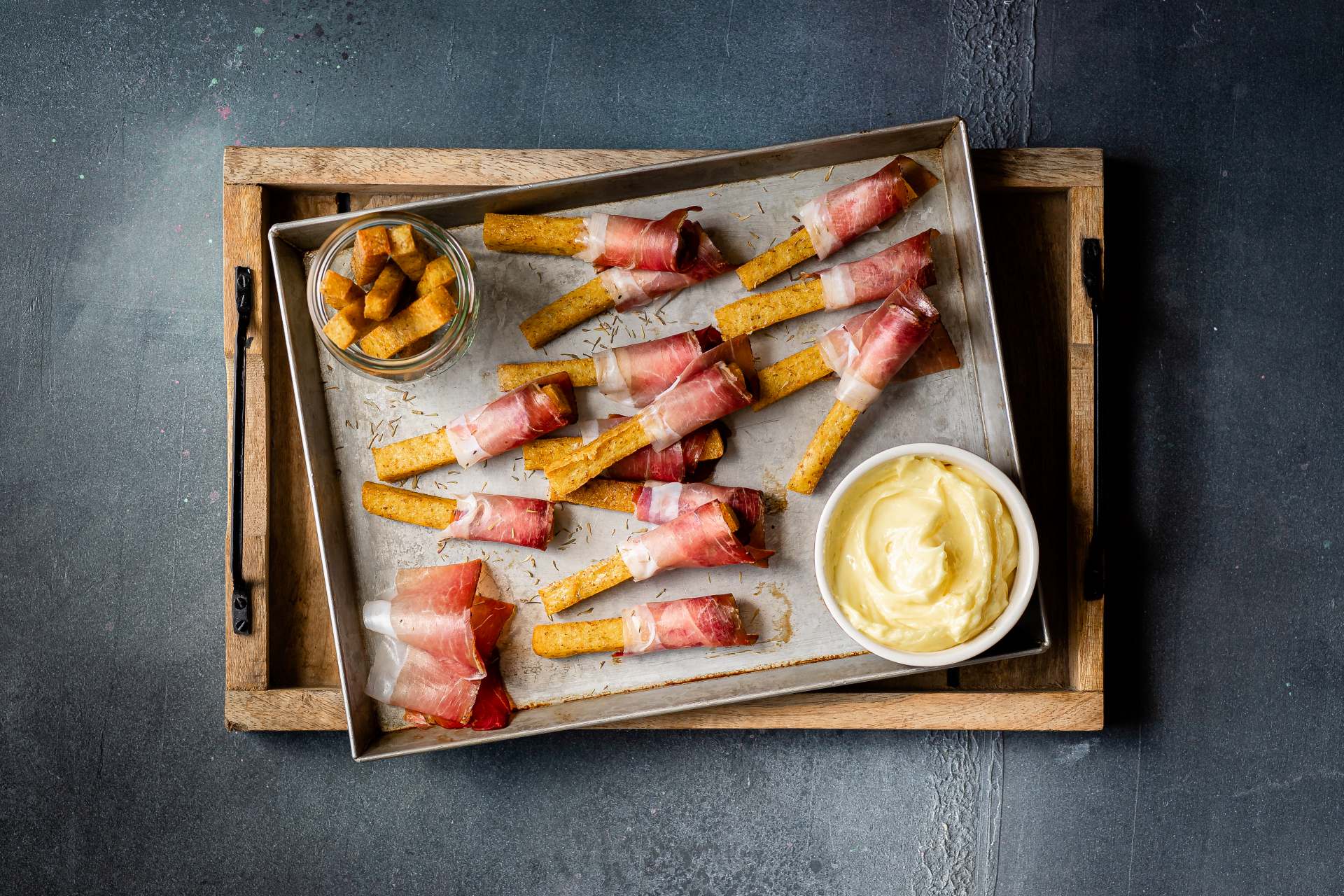 Knusprige Polenta-Chips, umhüllt von Südtiroler Speck g.g.A.-Scheiben, arrangiert auf einem Backblech mit Schälchen Aioli und Polenta-Würfeln im Hintergrund.