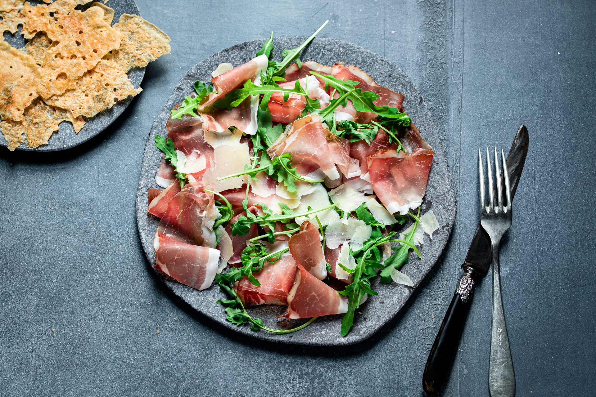 Südtiroler Speck g.g.A. mit frischer Rauke und Grana-Späne; daneben knusprige Roggenwaffel.
