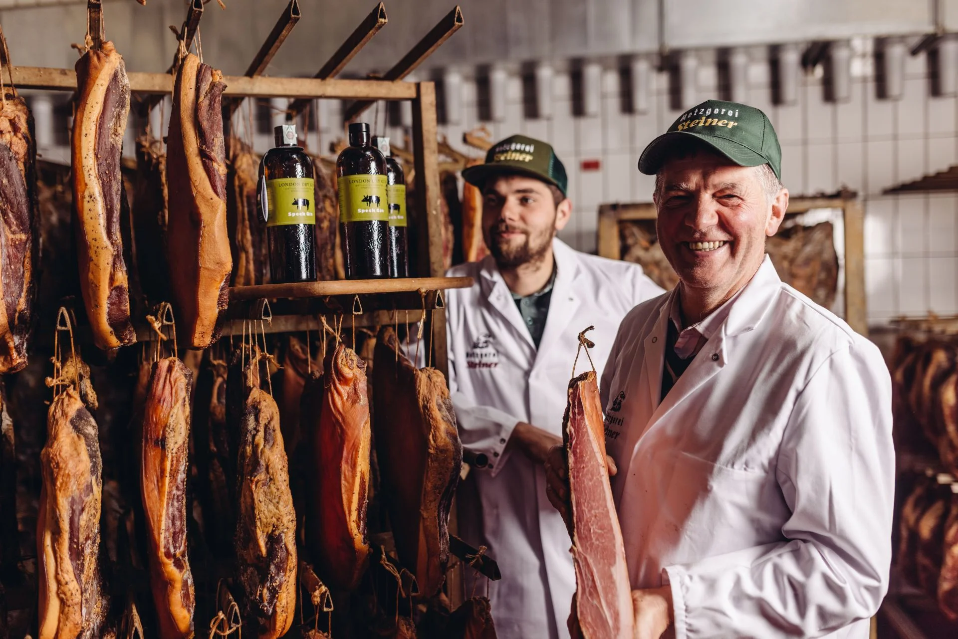 Produzenten Steiner im Reiferaum mit aufgehängten Stücken Südtiroler Speck g.g.A., einer hält ein Stück