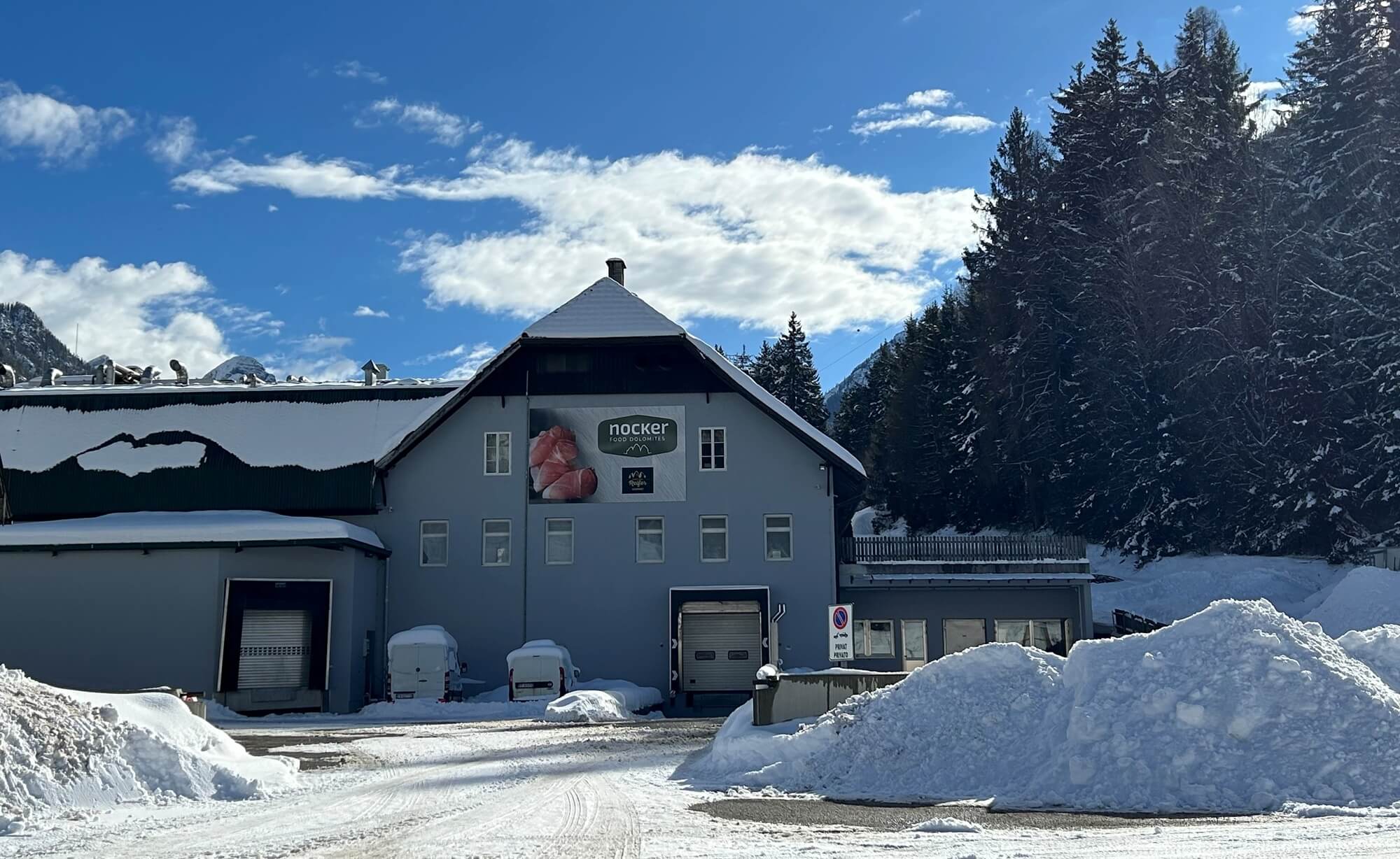 Edificio della sede Nocker in inverno con piazzale innevato e alberi davanti a paesaggio montano