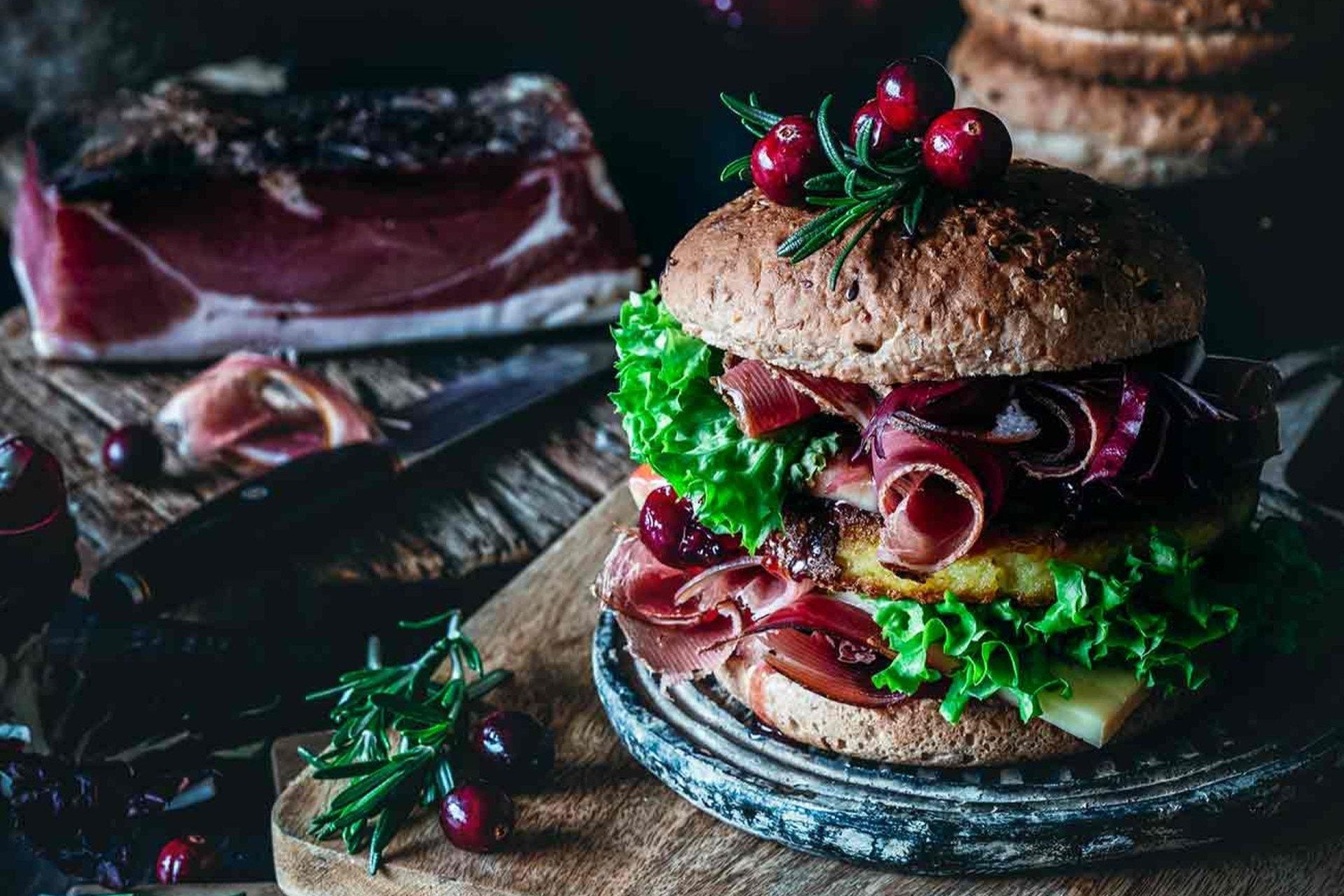 Burger rustique au pain complet avec speck, fromage fondu, salade verte et oignons, garni de canneberges et de romarin sur une planche en bois.