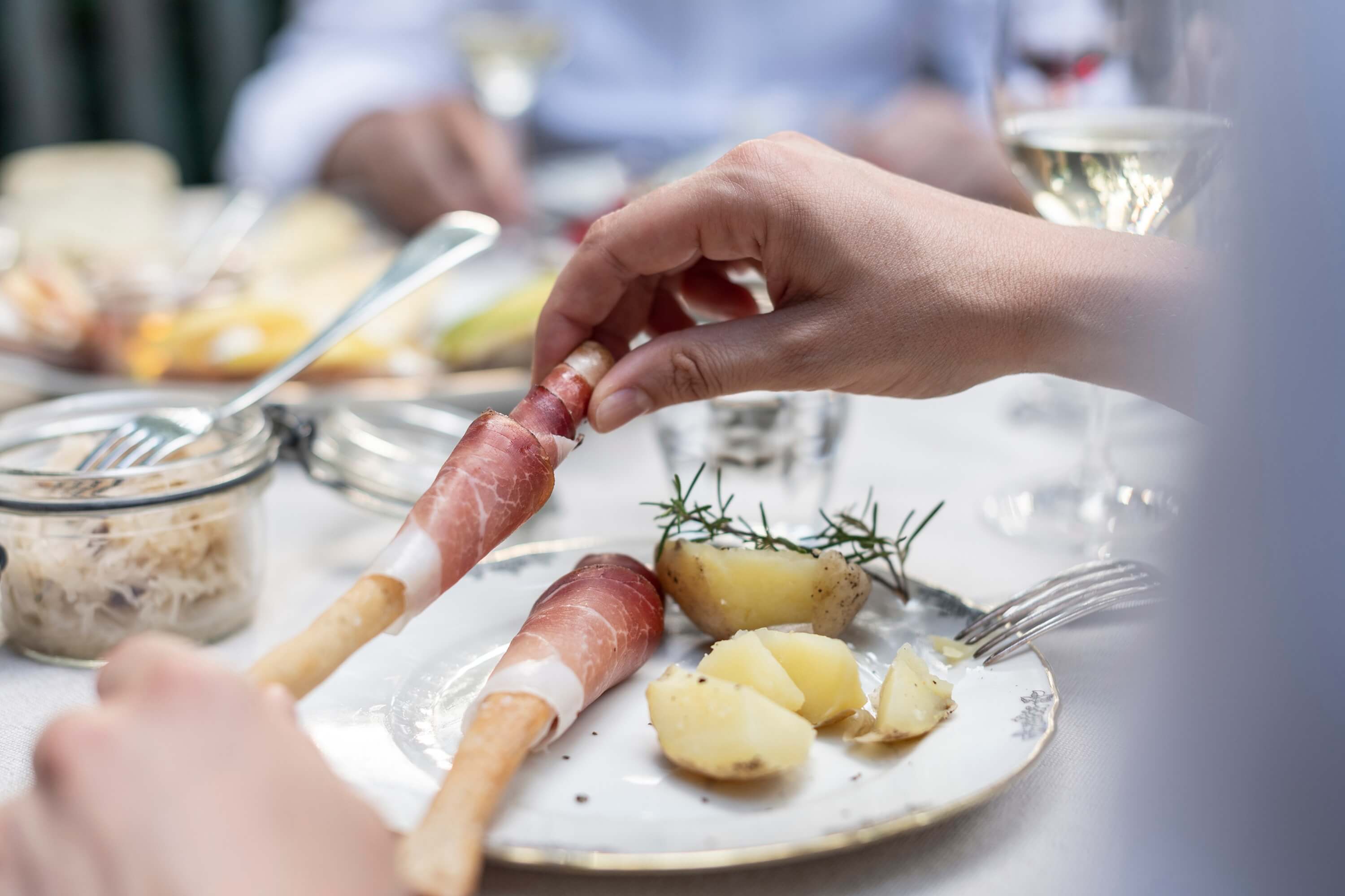 Hand rollt Speck Alto Adige g.g.A. um Grissini auf Teller mit Kartoffeln und Rosmarin.