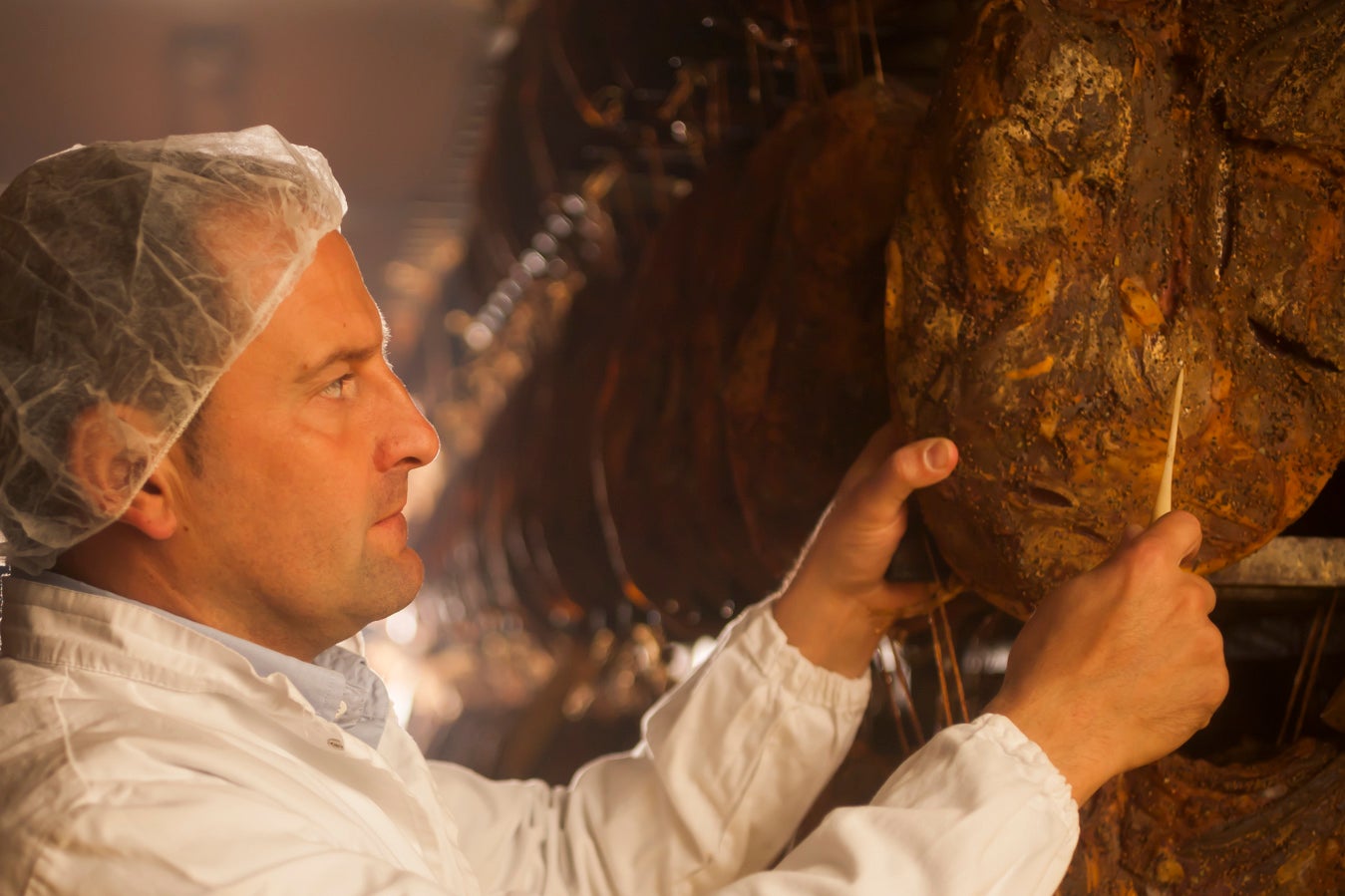 Hersteller mit Kittel und Haube prüft Südtiroler Speck g.g.A. mit Nadel während der Reifung in der Produktionstätte