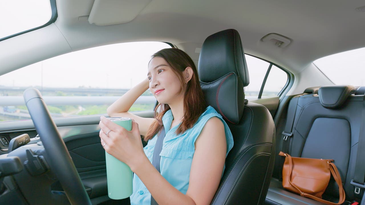 A lady sits in a self-driving car day dreaming and holding a flask of coffee