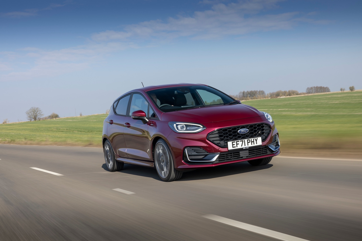 Maroon Ford Fiesta driving against a green field and blue skies