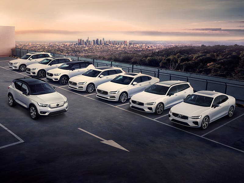 Row of various white Volvo models parked on roof top, with cityscape visible in the background