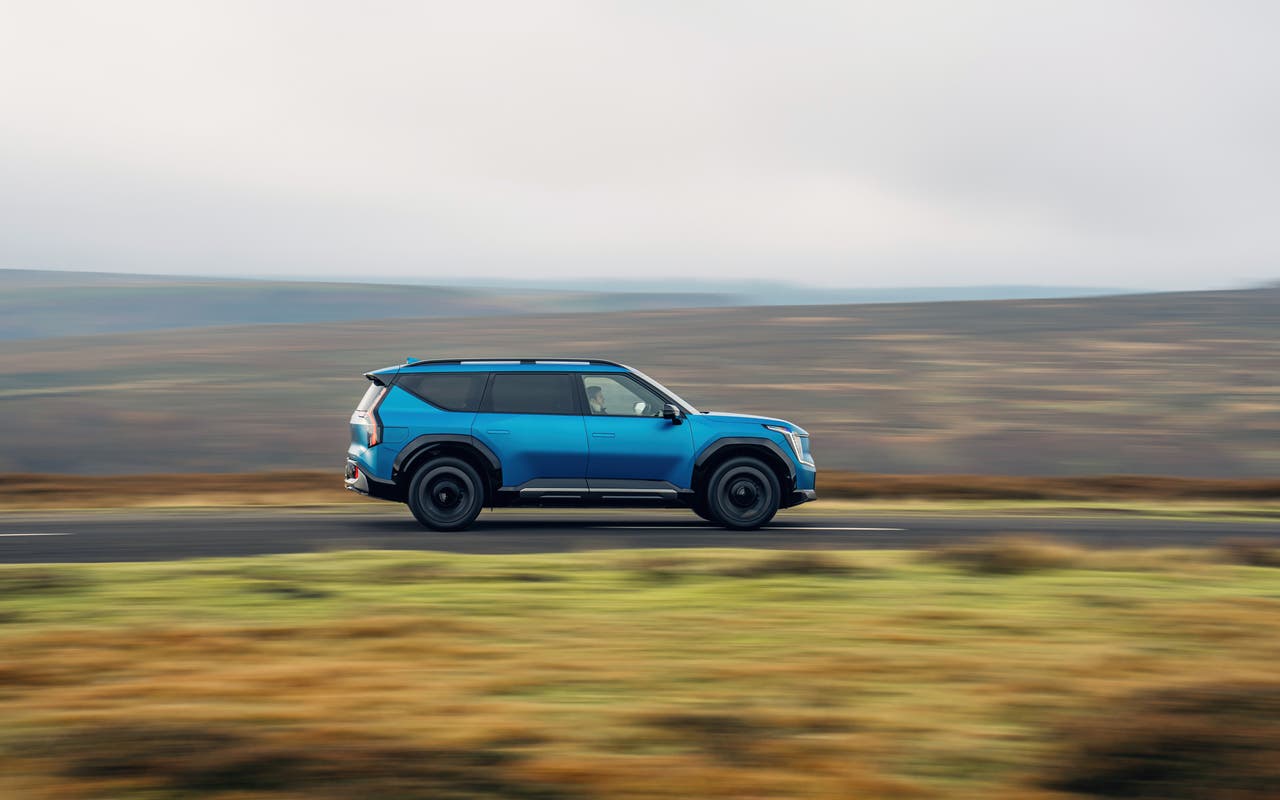 Blue Kia EV9 drives across a moorland road, viewed from the side