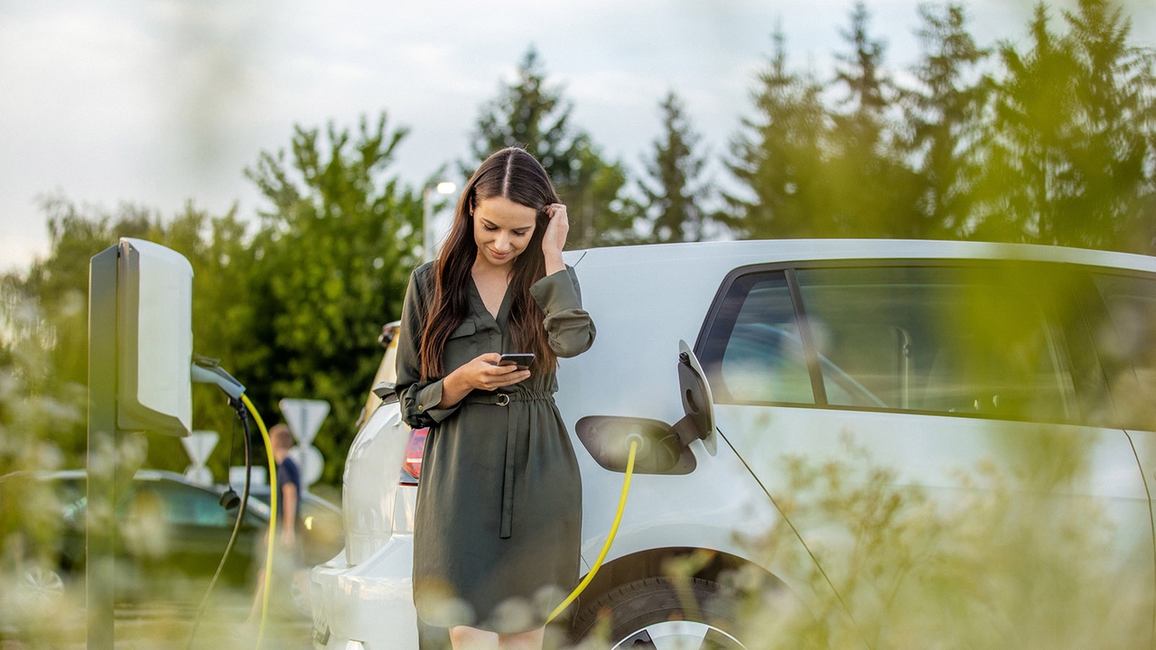 Lady stood next to a charging electric car. She's looking at her phone, presumably using an EV route planner to illustrate the writer's point.