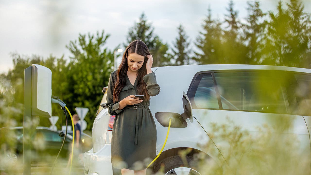 Lady stood next to a charging electric car. She's looking at her phone, presumably using an EV route planner to illustrate the writer's point.