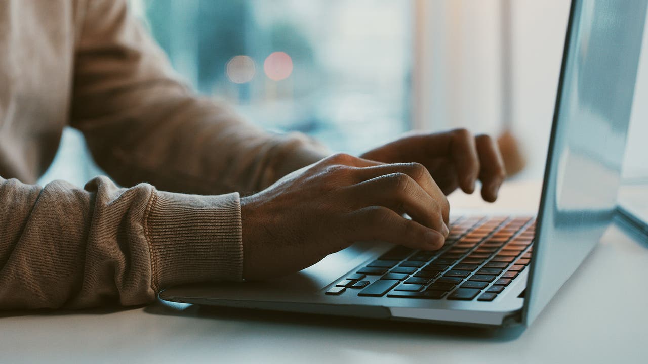 Disembodied hands type purposefully on a laptop, beige jumper