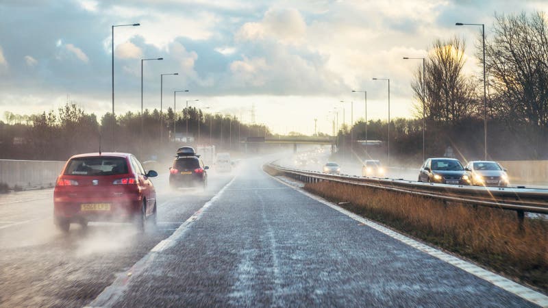 Point of view shot from the outside lane of a British motorway. There is dramatic spray as car tyres kick up rain from the damp road surface