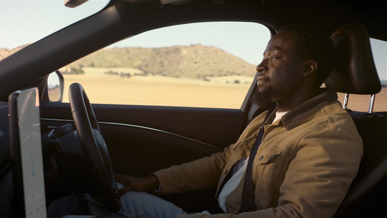 Man in camel-coloured coat sits behind the wheel of a semi-autonomous Ford Mustang Mach-E with his eyes on the road but hands off the wheel.