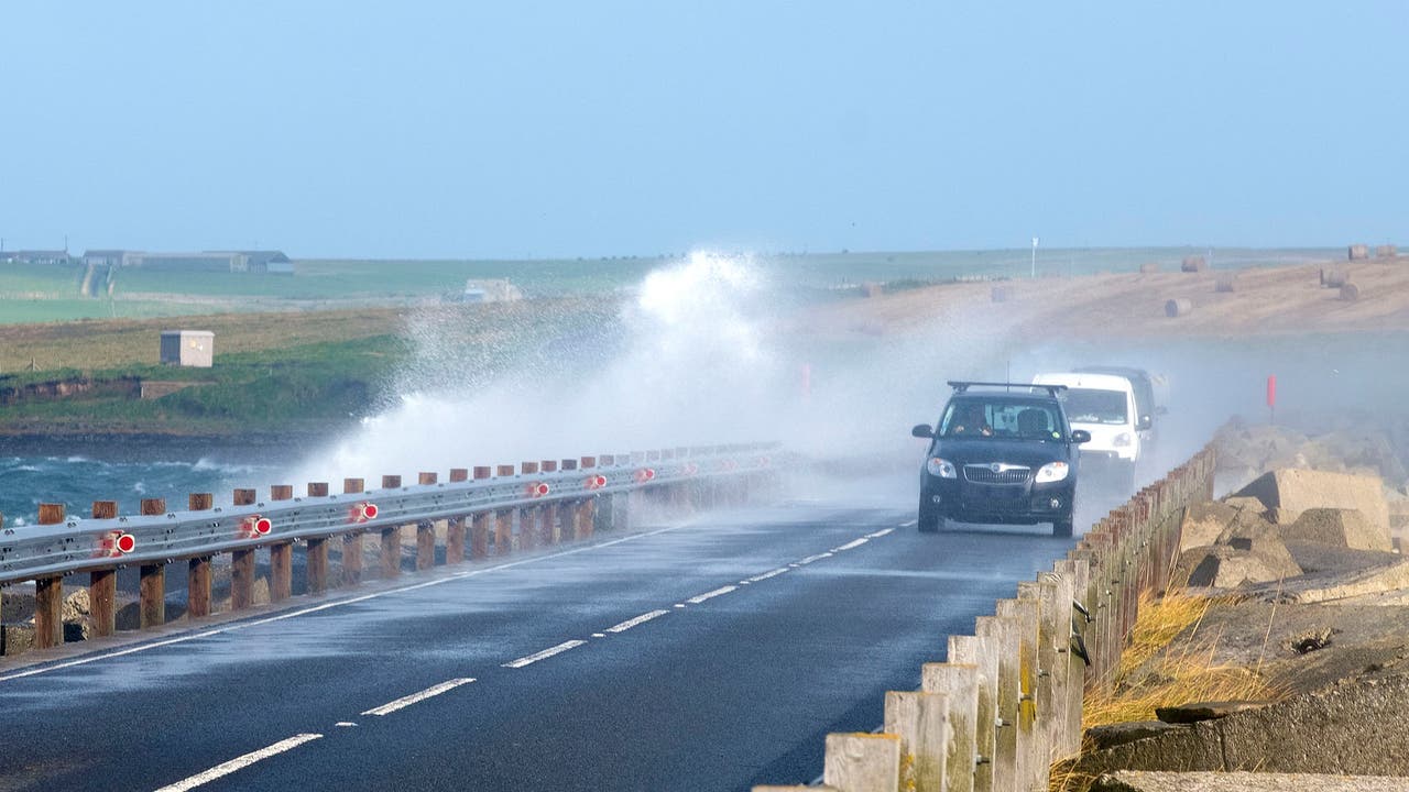 Vehicles driving on road next to water in windy conditions