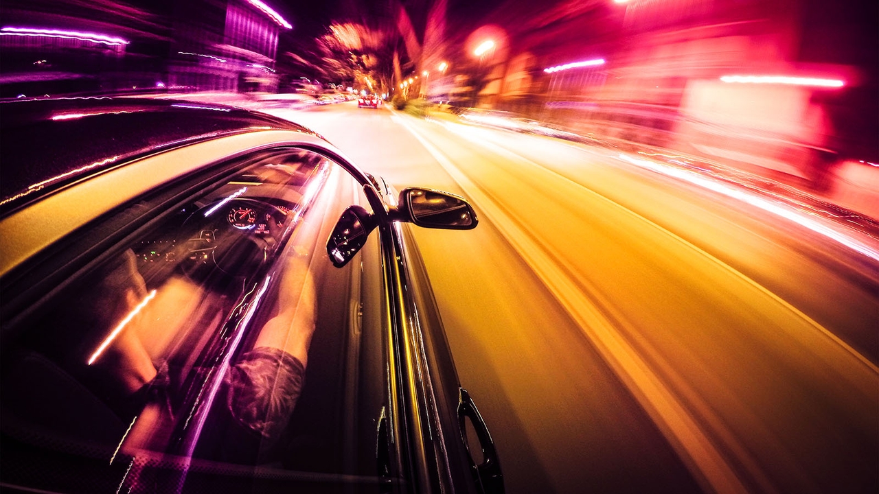 Shot of man driving a car down a road with neon lighting - the image is blurred as if to convey the idea of high speeds