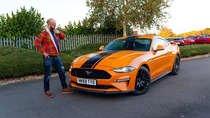 A handsome bald man scratches his chin while looking at a bright orange Ford Mustang