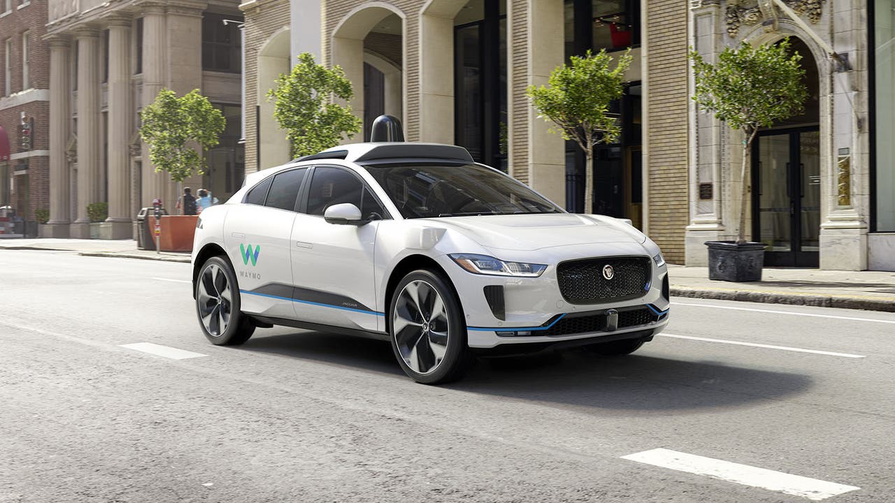 A white Jaguar I-Pace drives along equipped with Waymo self-driving equipment including a prominent LiDAR dome on the roof