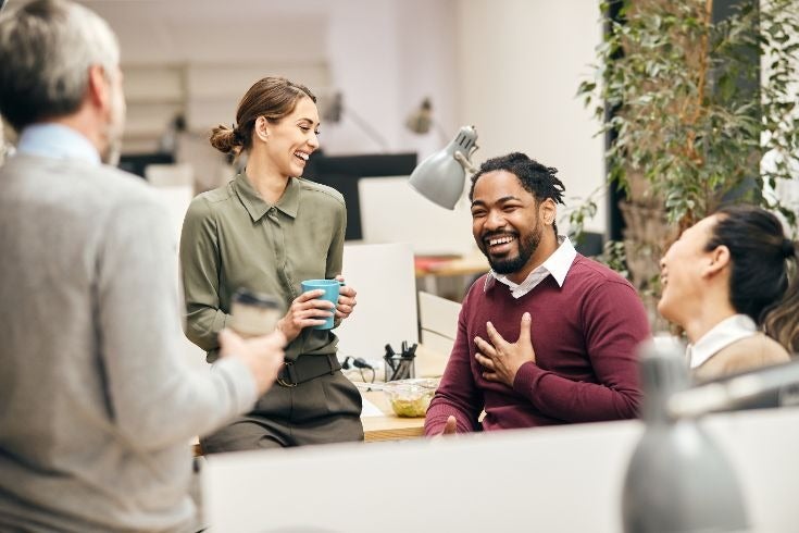 Colleagues standing around a desk in an office, holding drinks and talking, with desks and plants visible in the background.