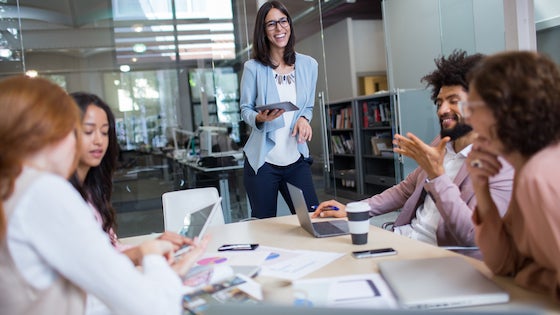 Colleagues in an office meeting room, with one person standing and holding a tablet while others look at their laptops.