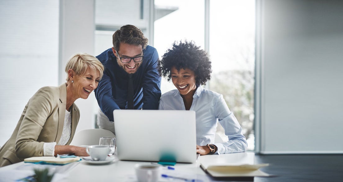 Three people gathered around a laptop at a desk in a bright office.