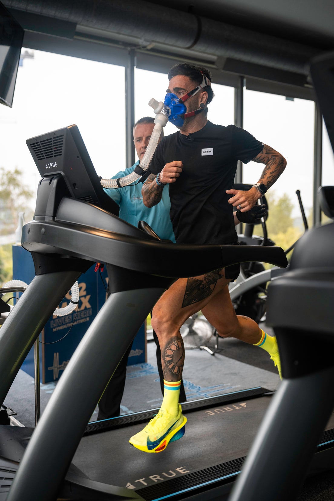 A man on an exercise bike with an oxygen mask on his face hooked up to a nearby monitor. A woman in a lab coat with a clipboard is taking notes next to him.