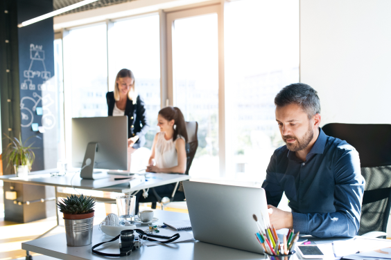 Man at his desk working on his laptop in the office, while his colleagues chat in the background.
